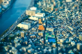 A vibrant cityscape with a blend of modern and traditional buildings is seen from above, featuring a distinctive orange building at the center. A river runs alongside the city with multiple bridges crossing it, while roads filled with vehicles connect various parts of the urban area. The tilt-shift effect creates a miniature model appearance, emphasizing a bustling yet organized city environment.