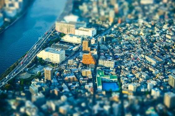 A vibrant cityscape with a blend of modern and traditional buildings is seen from above, featuring a distinctive orange building at the center. A river runs alongside the city with multiple bridges crossing it, while roads filled with vehicles connect various parts of the urban area. The tilt-shift effect creates a miniature model appearance, emphasizing a bustling yet organized city environment.