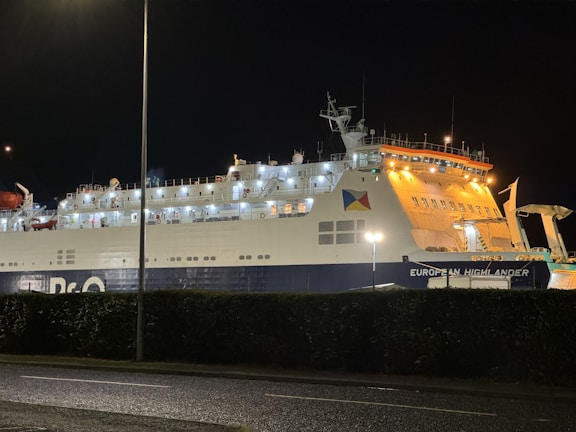 A large ferry with bright lights illuminating its side is docked at night. The ferry has a distinctive logo and is named 'European Highlander'. It is positioned beside a roadway, with a hedge partially obscuring the lower section of the hull.