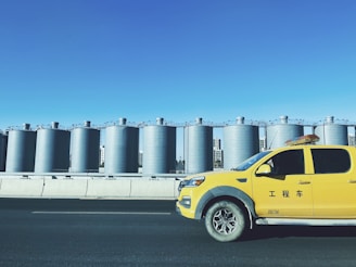 A row of large industrial silos with a bright blue sky in the background and a yellow pickup truck driving on a road in the foreground. The truck has characters written on its side.