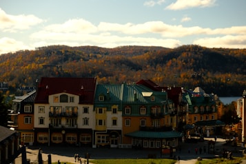 Colorful European-style buildings are nestled at the base of a tree-covered hill during autumn, with vibrant red, green, and yellow roofs and facades. The background includes rolling hills covered with fall foliage in hues of orange, yellow, and brown under a sky with scattered clouds. People can be seen walking on the street below the buildings.