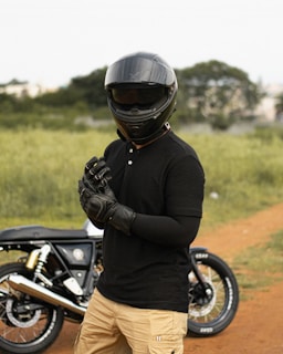 A person wearing a black helmet and gloves stands in an outdoor setting with a motorcycle in the background. The person is dressed in a black shirt and beige pants, and the setting appears to be a grassy area with a dirt path.