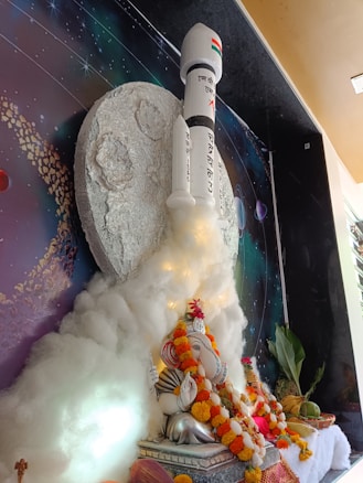 A decorative scene featuring a large model of a rocket launching into space, surrounded by artificial clouds. In front of the rocket sits a statue of Lord Ganesha adorned with marigold garlands, placed on a platform. The backdrop portrays outer space with celestial details, including a depiction of the moon and planets.