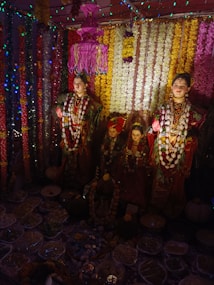 A vibrant scene with elaborately dressed statues adorned with floral garlands and jewelry. The backdrop is decorated with colorful floral arrangements in various hues. Illuminated by multicolored lights, the atmosphere is festive. Various offerings, including food items, are placed at the base.