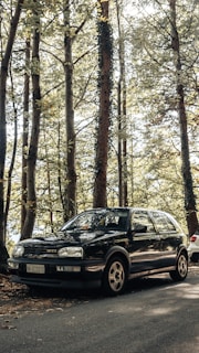 A black Volkswagen Golf GTI is parked on a road surrounded by tall trees with dense foliage. Sunlight filters through the leaves, casting dappled shadows on the car and ground.