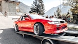 A bright red sports car is secured on a flatbed trailer, possibly being transported or showcased. Surrounding the car are several people, including some sitting on a bench and a cyclist passing by. The setting is outdoors with mountainous scenery in the background and some trees and a building visible.