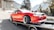 A bright red sports car is secured on a flatbed trailer, possibly being transported or showcased. Surrounding the car are several people, including some sitting on a bench and a cyclist passing by. The setting is outdoors with mountainous scenery in the background and some trees and a building visible.