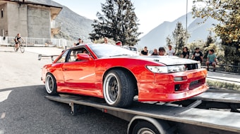 A bright red sports car is secured on a flatbed trailer, possibly being transported or showcased. Surrounding the car are several people, including some sitting on a bench and a cyclist passing by. The setting is outdoors with mountainous scenery in the background and some trees and a building visible.