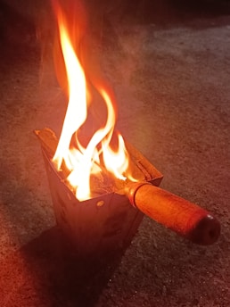 A metal container with a wooden handle is holding a small fire, with bright orange and yellow flames rising into the air. The surface under the container is rough and appears to be concrete.