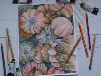 A watercolor painting of various pumpkins displayed on a white wooden surface. Surrounding the painting are colored pencils, paint tubes, a paintbrush, and pages from a book.