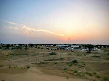 A vast desert landscape is depicted with a few scattered bushes and patches of greenery. In the distance, numerous white tents are lined up, suggesting a campsite or festival area. Several vehicles are parked nearby, indicating human activity. The sky is a soft gradient as the sun begins to set, casting a warm glow across the scene.