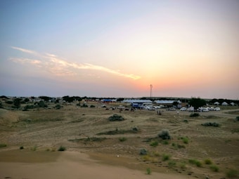 A vast desert landscape is depicted with a few scattered bushes and patches of greenery. In the distance, numerous white tents are lined up, suggesting a campsite or festival area. Several vehicles are parked nearby, indicating human activity. The sky is a soft gradient as the sun begins to set, casting a warm glow across the scene.