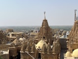 An architectural landscape featuring intricately designed, historic sandstone buildings with ornate towers and domes. The structures exhibit traditional craftsmanship with detailed carvings, set against a vast, open background under a clear blue sky.