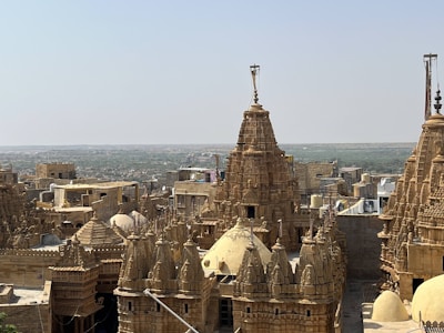 An architectural landscape featuring intricately designed, historic sandstone buildings with ornate towers and domes. The structures exhibit traditional craftsmanship with detailed carvings, set against a vast, open background under a clear blue sky.
