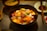 A bowl of colorful seafood and garnishes, including salmon, tuna, white fish, and roe, topped with herbs and a small dollop of wasabi. The bowl is placed on a textured table mat with a wooden spoon nearby, and a cup of soup and a small dish of sauce in the background.