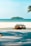 A scenic beach setting featuring a pathway leading to a sandy shore with clear blue skies and an emerald green ocean. A small island is visible in the distance. In the foreground, there is a retro-styled bus partially hidden behind a large rock, with a palm tree branch hanging over the scene.
