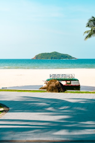 A scenic beach setting featuring a pathway leading to a sandy shore with clear blue skies and an emerald green ocean. A small island is visible in the distance. In the foreground, there is a retro-styled bus partially hidden behind a large rock, with a palm tree branch hanging over the scene.