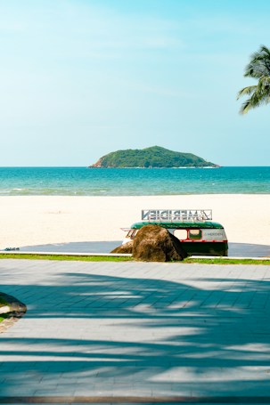 A scenic beach setting featuring a pathway leading to a sandy shore with clear blue skies and an emerald green ocean. A small island is visible in the distance. In the foreground, there is a retro-styled bus partially hidden behind a large rock, with a palm tree branch hanging over the scene.
