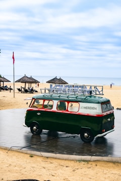A vintage green van with red accents and the Le Meridien logo is parked on a sandy beach near the ocean. It's surrounded by a stone platform and nearby are several thatched-roof umbrellas with sun loungers. In the background, people are visible enjoying the beach, and a red flag is flying in the breeze. The sky is cloudy yet bright, creating a serene atmosphere.