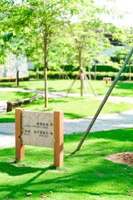 A signpost stands prominently on a well-manicured green lawn, with two wooden posts supporting a stone plaque directing towards a lagoon swimming pool, beach, and horizon. The background features lush green trees and a paved pathway suggesting a park or resort setting.