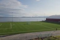A coastal soccer field with a bright green artificial turf is situated near a large body of water. In the background, distant mountains are visible beneath a partly cloudy sky. A large, red building with a dome-like roof is positioned to the right. The area is enclosed by a metal fence, and a person can be seen walking along the perimeter.