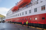 A large passenger ship docked at a port with people cleaning its side. The ship has a distinct red, white, and black color scheme, with large orange lifeboats mounted on its side. Several workers in high-visibility clothing are using long poles to clean the windows and sides of the ship. The setting appears to be a harbor with a hillside town visible in the background.