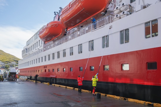 A large passenger ship docked at a port with people cleaning its side. The ship has a distinct red, white, and black color scheme, with large orange lifeboats mounted on its side. Several workers in high-visibility clothing are using long poles to clean the windows and sides of the ship. The setting appears to be a harbor with a hillside town visible in the background.