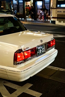 A white sedan with a Japanese license plate is parked on the street. The background shows an urban setting at night with blurred figures of people and a bus stop. The car's brake lights are illuminated, adding a warm glow to the scene.