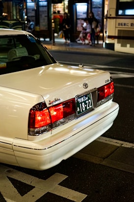 A white sedan with a Japanese license plate is parked on the street. The background shows an urban setting at night with blurred figures of people and a bus stop. The car's brake lights are illuminated, adding a warm glow to the scene.
