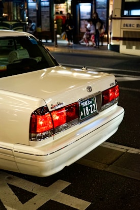 A white sedan with a Japanese license plate is parked on the street. The background shows an urban setting at night with blurred figures of people and a bus stop. The car's brake lights are illuminated, adding a warm glow to the scene.