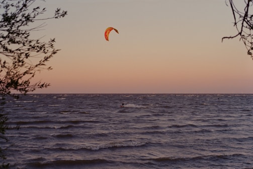 Sunset silhouette of a kite surfer gliding over calm sea with vibrant orange and pink sky.