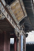 An intricately carved wooden sign with Chinese characters is mounted on a traditional building with ornate architectural details. The structure features wooden beams, decorated with elaborate carvings and faded paint, suggesting historical significance. Roof tiles and columns are visible, enhancing the traditional aesthetic.