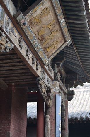 An intricately carved wooden sign with Chinese characters is mounted on a traditional building with ornate architectural details. The structure features wooden beams, decorated with elaborate carvings and faded paint, suggesting historical significance. Roof tiles and columns are visible, enhancing the traditional aesthetic.