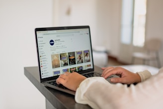 A person is using a laptop on a dark-colored desk, viewing a TikTok profile. The screen displays various video thumbnails and interface elements. The setting is a bright, modern room with minimal furniture in the background.