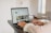 A person is using a laptop on a dark-colored desk, viewing a TikTok profile. The screen displays various video thumbnails and interface elements. The setting is a bright, modern room with minimal furniture in the background.