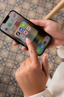 A person is holding a smartphone, displaying a folder of social media apps including TikTok, LinkedIn, WhatsApp, Pinterest, YouTube, and Snapchat. The background consists of a patterned floor.