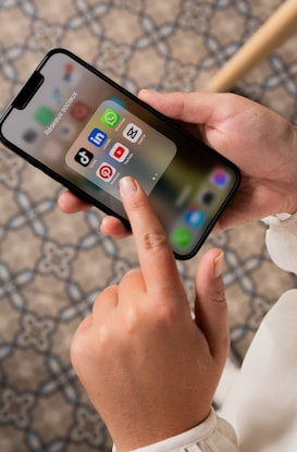 A person is holding a smartphone, displaying a folder of social media apps including TikTok, LinkedIn, WhatsApp, Pinterest, YouTube, and Snapchat. The background consists of a patterned floor.