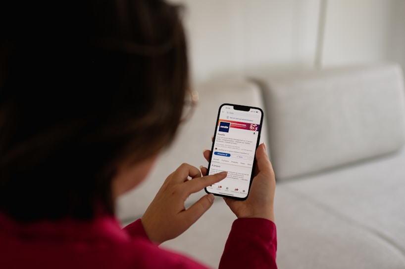 A person is holding a smartphone, engaging with an app that resembles LinkedIn. The individual is using their thumb to scroll or select on the screen. They are wearing a red top, and the background includes a light-colored couch.