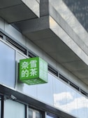 A modern building exterior featuring a prominent green sign with white Chinese characters, mounted on a shiny facade of glass and concrete. The sign is a cube shape extending from the building, reflecting some light due to its glossy surface.