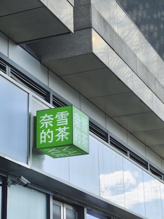 A modern building exterior featuring a prominent green sign with white Chinese characters, mounted on a shiny facade of glass and concrete. The sign is a cube shape extending from the building, reflecting some light due to its glossy surface.