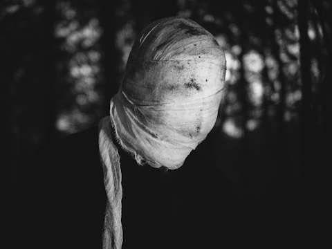 A figure with their head completely wrapped in a white cloth, standing against a dark, blurred background. The cloth appears dirty and loosely tied, conveying an eerie, mysterious atmosphere.