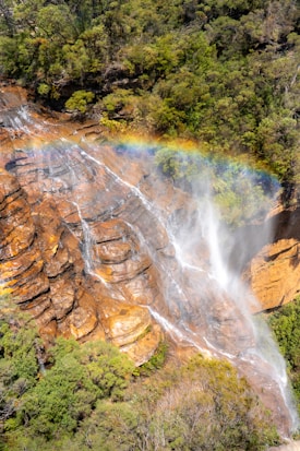 A vibrant rainbow curves over a cascading waterfall surrounded by lush greenery on rocky cliffs. The water flows energetically down the reddish-brown rock face, creating a mist that enhances the colorful arc above.