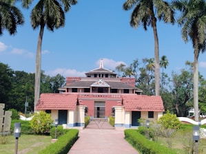 A large heritage-style building with a red-tiled roof and intricate architectural details, surrounded by tall palm trees and lush greenery. The structure is accessed via a paved pathway lined with manicured hedges.