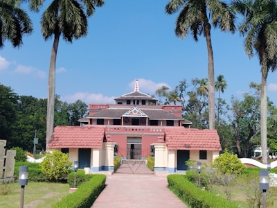 A large heritage-style building with a red-tiled roof and intricate architectural details, surrounded by tall palm trees and lush greenery. The structure is accessed via a paved pathway lined with manicured hedges.