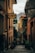 A narrow, picturesque alleyway flanked by colorful buildings with shuttered windows. There are signs for restaurants along the walls, and potted plants add greenery. The alley gently slopes down with cobblestone steps and leads to a scenic view of distant mountains.