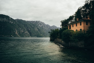 A serene lakeside scene with a large, peach-colored house nestled among dense green foliage on the right. Mountains rise majestically in the background under a cloudy sky. The water is calm, reflecting the surrounding landscape.