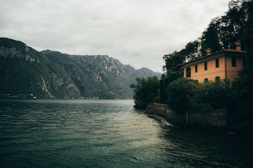 A serene lakeside scene with a large, peach-colored house nestled among dense green foliage on the right. Mountains rise majestically in the background under a cloudy sky. The water is calm, reflecting the surrounding landscape.