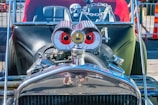 A customized car with a vintage design features eye-catching modifications including large cartoonish red eyes on the engine, a skull decoration on the dashboard, and a metallic chain. The bright and shiny chrome parts reflect sunlight, giving it a polished look.