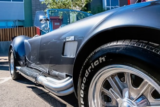 A glossy, vintage sports car is seen with a shiny metallic body and chrome details. The perspective focuses closely on the side and back tire, highlighting its polished surface and distinctive side exhaust pipes. The car is parked on an asphalt surface with modern buildings and a fenced area in the background.