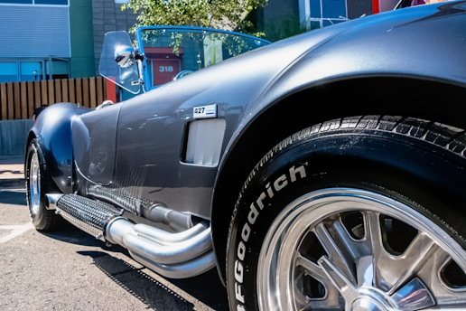 A glossy, vintage sports car is seen with a shiny metallic body and chrome details. The perspective focuses closely on the side and back tire, highlighting its polished surface and distinctive side exhaust pipes. The car is parked on an asphalt surface with modern buildings and a fenced area in the background.
